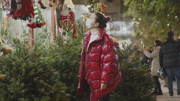 Caucasian young lady in big red puffer jacket and coronavirus face mask in the Christmas market. Cowboy shot. Stok Resim