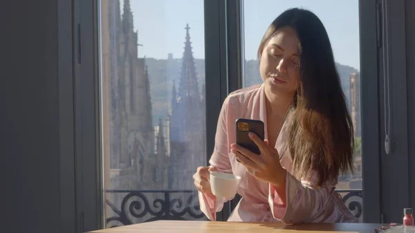 Jovem mista de raça feminina em um robe de seda rosa, com um smartphone na mão está bebendo sua primeira xícara de café com vistas Barcelona sobre o fundo. Retrato de tiro médio de alta qualidade Imagens De Bancos De Imagens