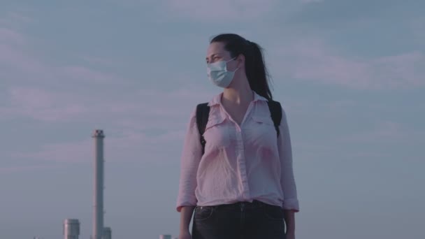Pagi atau malam di alun-alun kosong seorang wanita muda kaukasia berdiri di topeng medis dengan latar belakang pabrik perkotaan — Stok Video