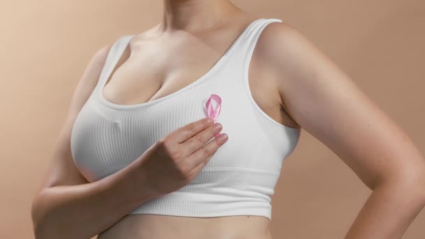 Кавказька жінка в білому безшовному бюстгальтерці поклала рожеву стрічку на рак молочної залози. Студійні анонімні середні зйомки на бежевому тлі. — стокове відео