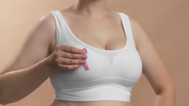 Junge Kaukasierin in weißem, nahtlosem BH mit rosa Schleife gegen Brustkrebs. Studio anonymes Medium aufgenommen auf beigem Hintergrund. — Stockvideo