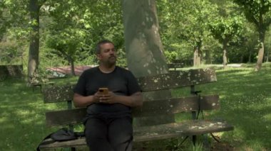 Orta yaşlı, melez bir yetişkin adam parkta ya da ormanda bir bankta oturup telefonunu izliyor. Yavaş çekim videosu.