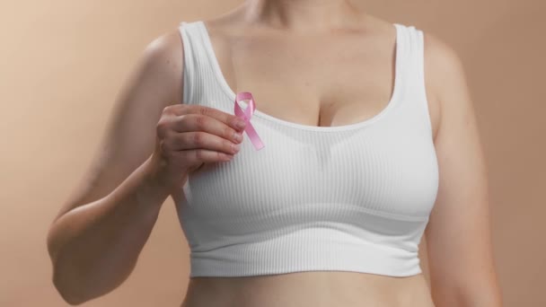 Caucasian woman in a white seamless bra put pink ribbon for breast cancer. Studio anonymous medium shot on beige background. — Stockvideo