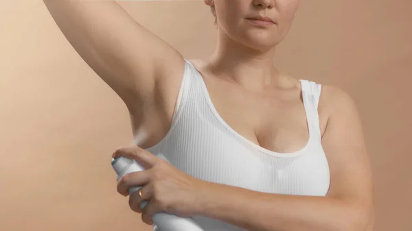 Caucasian anonymous girl in white top with a hand spray deodorant on her underarm. Studio photo image on beige background. — Stock Photo, Image