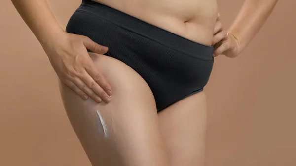 Eine vollbusige Kaukasierin in schwarzer Unterwäsche cremt ihr Bein ein, um es zu massieren. Beige Studio Hintergrund qualitativ hochwertige Foto. — Stockfoto
