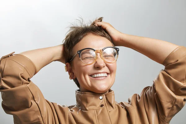 Glad tjej som rör vid huvudet. Överlycklig kaukasiska kvinna med kort frisyr i glasögon och beige jacka leende. Porträtt på vit bakgrund foto bild. — Stockfoto