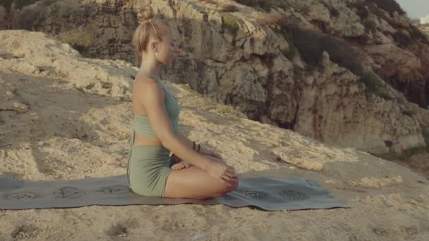 Blond young caucasian female yoga teacher meditating in the mountains near the sea. Panned shot slow motion high quality video footage. — Stock Video