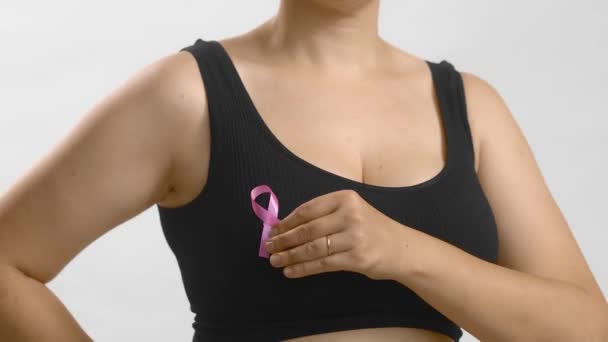 Caucasian woman in a black seamless bra put with her hand pink ribbon for breast cancer. Studio anonymous medium shot on white background. — Stock Video