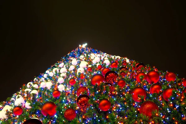 Christmas tree decorated with blue, white and red balls, and electric garlands against the black night sky, view from below