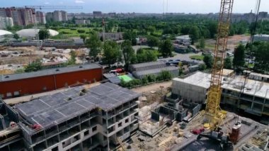 The camera moves from top to bottom, revealing a construction site, an unfinished building and a yellow tower crane on rails. In background you can see a green park and new buildings under a blue sky.