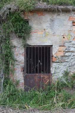 Paslı metal kapı ve parmaklıklı pencere. Parçalanmış tuğla duvarda zincir var..