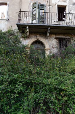 Çürüyen bir balkon, paslı bir korkuluk ve kısmen açılmış ahşap kapılar..