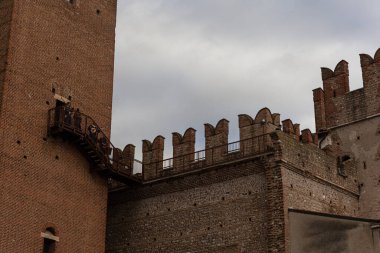 Eski bir kırmızı tuğladan kale ya da kale manzarası, iki kuleyi birbirine bağlayan çentikli siperler ve küçük ahşap bir köprü veya balkon gösteriyor..
