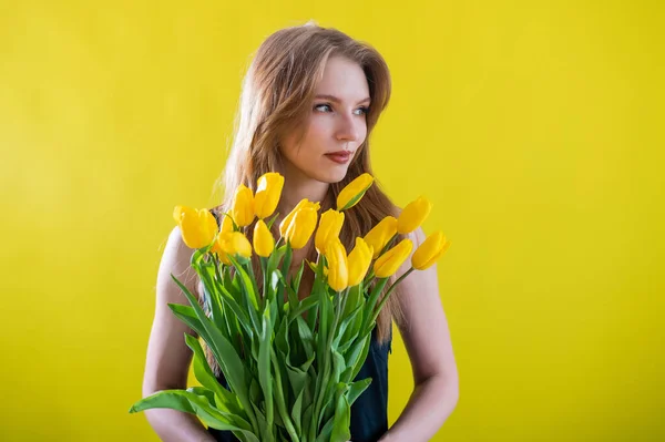 Kaukasische Frau mit einem Arm voller gelber Tulpen auf gelbem Hintergrund. Internationaler Frauentag. Strauß Frühlingsblumen — Stockfoto