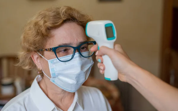 Um médico mede a temperatura de uma mulher idosa usando uma máscara com um termômetro infravermelho. visita domiciliar check-up médico — Fotografia de Stock