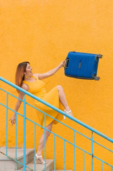 Schöne rothaarige lächelnde Frau kommt freudig die Treppe hinunter und schwenkt einen blauen Koffer auf gelbem Grund — Stockfoto