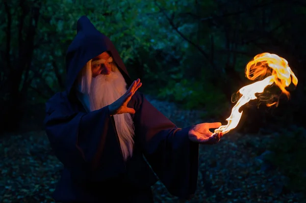 Yaşlı, kır saçlı bir cadı ormanda ateş büyüsü yapıyor. Büyücü kostümlü adam. — Stok fotoğraf