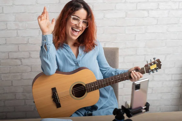 Een aantrekkelijke roodharige vrouw blogs op muziek. Het meisje zingt, speelt gitaar en neemt video op met een smartphone — Stockfoto
