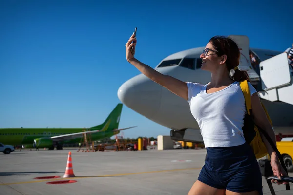 Kaukasische rothaarige Frau, die ein Selfie auf dem Hintergrund des Flugzeugs macht. Das Mädchen wird vor dem Flug auf dem Rollfeld fotografiert — Stockfoto