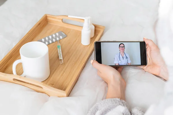 Μια γυναίκα βρίσκεται στο κρεβάτι με ένα κρύο και συμβουλεύεται έναν γιατρό σε απευθείας σύνδεση από το smartphone. Διαδικτυακή ιατρική. Ιός του κερατοειδούς — Φωτογραφία Αρχείου
