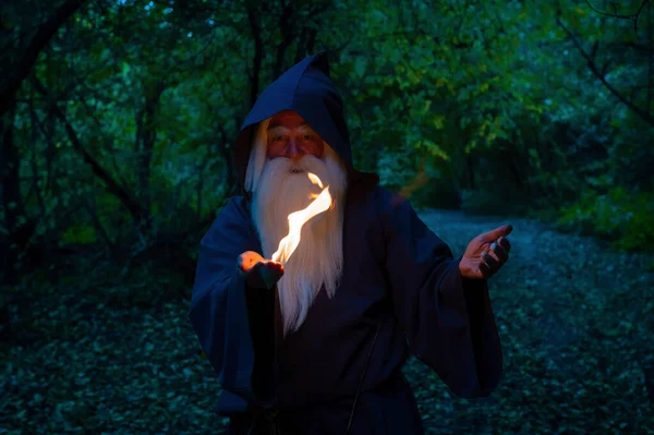 Yaşlı, kır saçlı bir cadı ormanda ateş büyüsü yapıyor. Büyücü kostümlü adam. — Stok fotoğraf