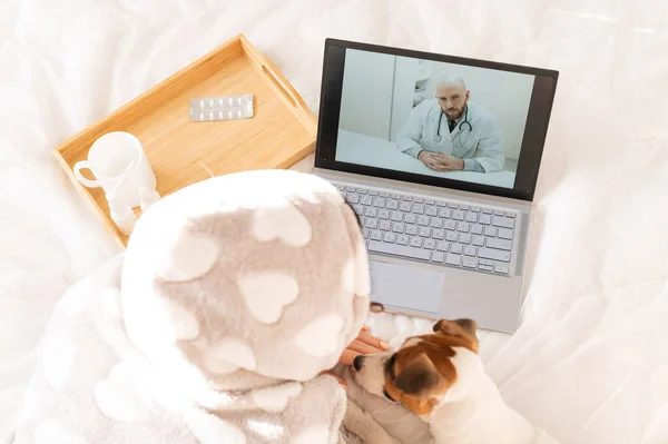 Хвора жінка лежить в ліжку і розмовляє з чоловіком-лікарем на ноутбуці. Віддалений прийом з терапевтом — стокове фото