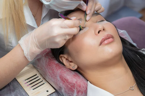 Close-up de uma bela mulher asiática no procedimento de extensão de pestanas. Cílios falsos de volume duplo. Lashmaker no trabalho. — Fotografia de Stock