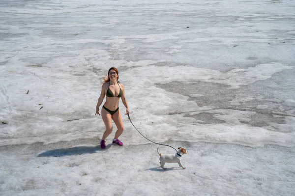 Rudowłosa kobieta w bikini spaceruje z psem na zaśnieżonej plaży — Zdjęcie stockowe