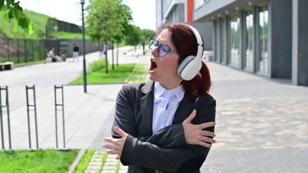 Mulher de negócios caucasiana em um terno ouvindo música com fones de ouvido e cantando ao ar livre — Vídeo de Stock