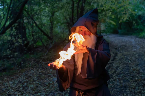 Un anciano brujo canoso lanza un hechizo de fuego en el bosque. Hombre en traje de mago — Foto de Stock