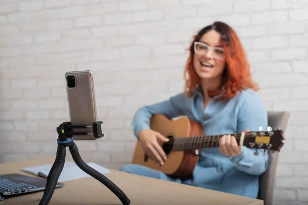 Egy nő online sugároz otthon a telefonján. A lány élőben énekel és gitározik. — Stock Fotó