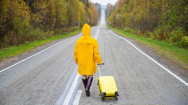 Une femme en imperméable jaune marche le long de la route avec une valise jaune en automne — Photo