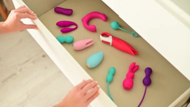 Bir kadın komodinin yanındaki masayı vibratör koleksiyonuyla kapatıyor.