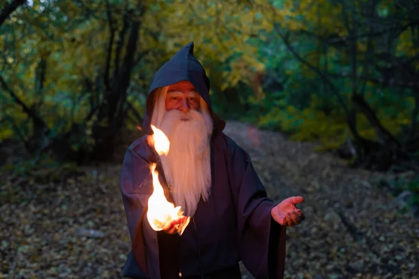 Een oudere grijsharige heksenmeester spreidt een vuurspreuk uit in het bos. Man in tovenaarskostuum — Stockfoto