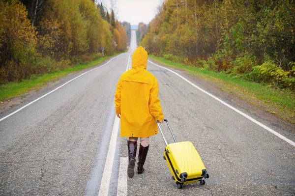 Une femme en imperméable jaune marche le long de la route avec une valise jaune en automne — Photo