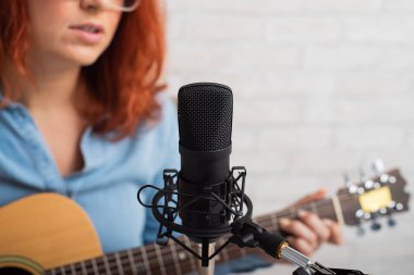 Kızıl saçlı bir kadın akustik gitar çalar ve kayıt stüdyosundaki bir mikrofona şarkı söyler.