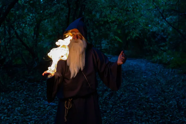 Un anciano brujo canoso lanza un hechizo de fuego en el bosque. Hombre en traje de mago — Foto de Stock
