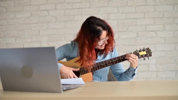 A nő megtanul gitározni a laptopon lévő videóból. Távolságtartó zene a karanténban — Stock videók