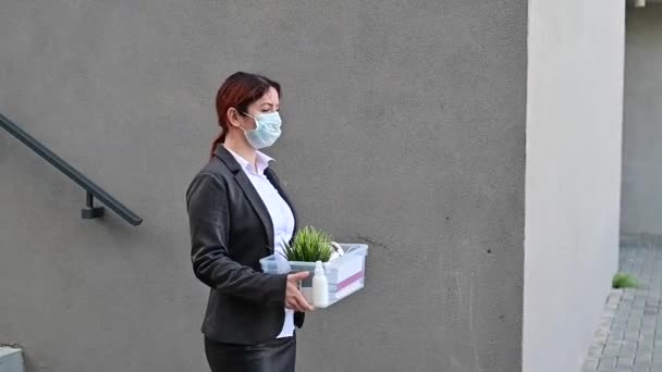 마스크를 쓴 불행 한 여성이 계단 뒤에 사적 인 물건들이 담긴 상자를 들고 거리를 걷고 있습니다. 한 여성 사무실 직원이 해고되었다. 유행병이 맹위를 떨치던 경제 위기 19. — 비디오