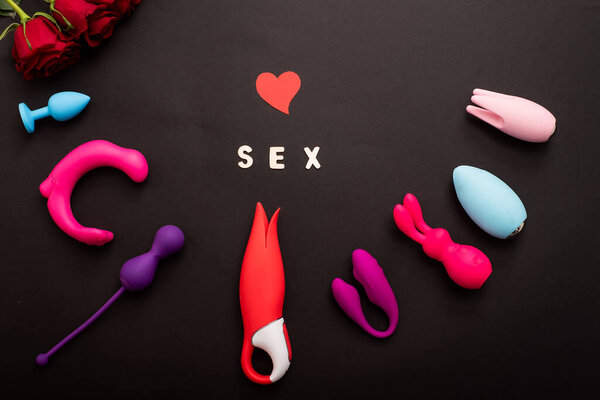The inscription sex is a heart of roses and a collection of vibrators on a black background. Sex toys of various shapes. Assortment of sex shop