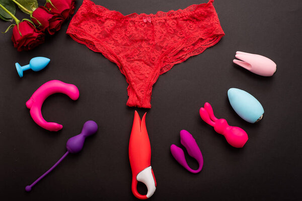 Top view of womens lace panties red roses and different sex toys. Collection of multicolored vibrators on a black background.