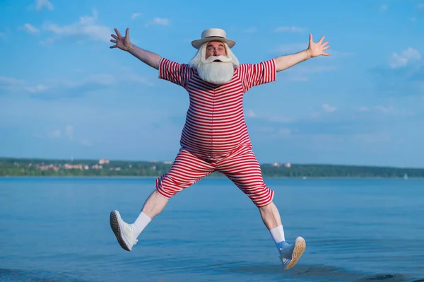 Un uomo anziano in costume da bagno retrò a righe corre e salta lungo la spiaggia. Un vecchio dai capelli grigi allegro con la barba in un cappello riposa al mare. — Foto Stock