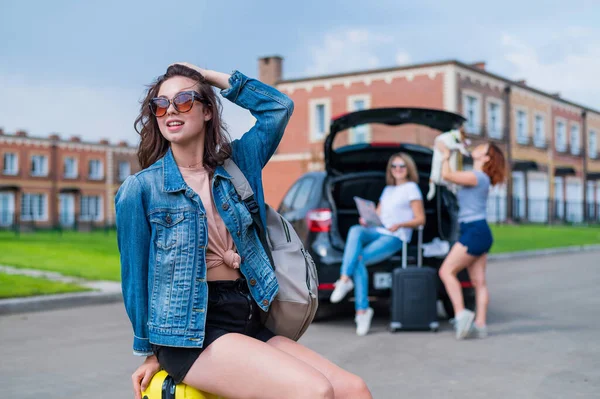 Drei Freundinnen gehen auf Roadtrip. Junge Frau posiert mit einem Koffer. Zwei Mädchen sitzen auf dem Kofferraum eines Autos und betrachten eine Landkarte. — Stockfoto