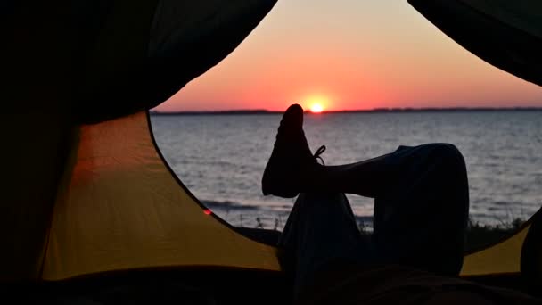 Vue des jambes féminines dans une tente touristique au coucher du soleil sur la plage. La femme fait de la randonnée et campe au bord de la rivière. — Video