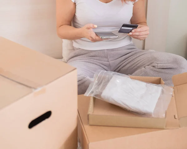Online shopping using the application on the phone. A faceless woman sits in bed unpacks an order and holds a smartphone and a credit card. — Stock Photo, Image