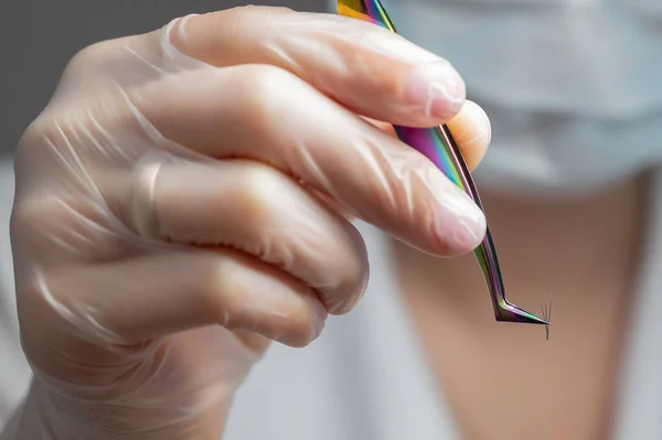 Lashmaker in een masker houdt een bos van wimpers met pincet. Close-up van een werktuig van een meester in een schoonheidssalon — Stockfoto