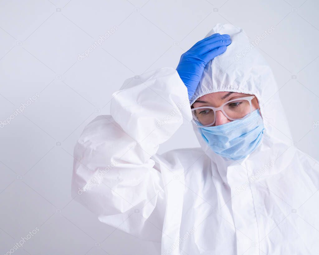 A woman in a protective suit and a medical mask is holding her head. Doctor virologist with problems at work