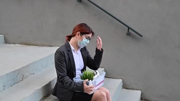 Уволенная женщина с коробкой личных вещей сидит на лестнице снаружи. Женщина в медицинской маске уволена из-за финансового кризиса в короновирусе. Безработица. — стоковое видео