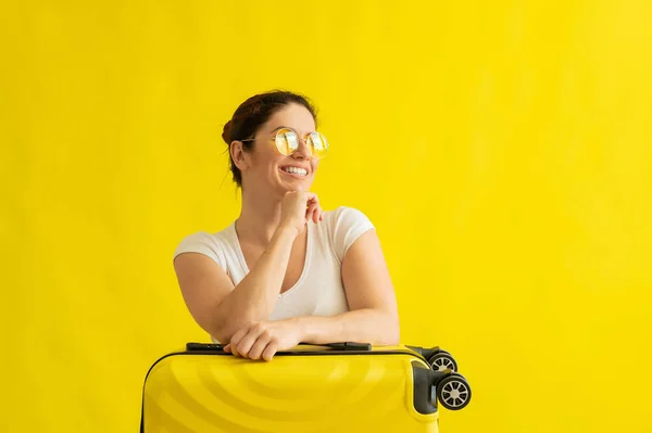 Kobieta z doskonałym śnieżnobiałym uśmiechem w okularach przeciwsłonecznych przytula walizkę na żółtym tle. Podekscytowana dziewczyna czeka na podróż na wakacjach i przytula się do torby. — Zdjęcie stockowe