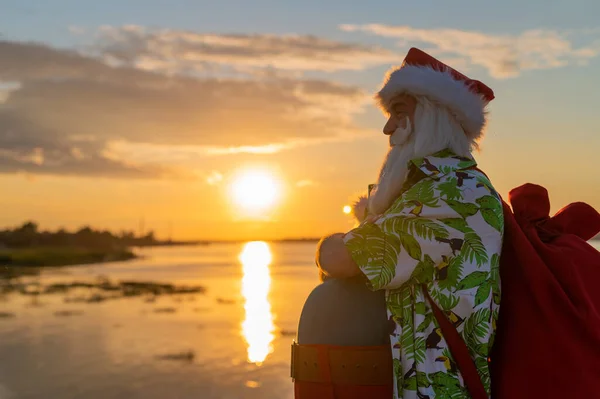 Papai Noel em shorts e uma camisa caminha ao longo da praia ao pôr do sol. Natal em um país quente — Fotografia de Stock